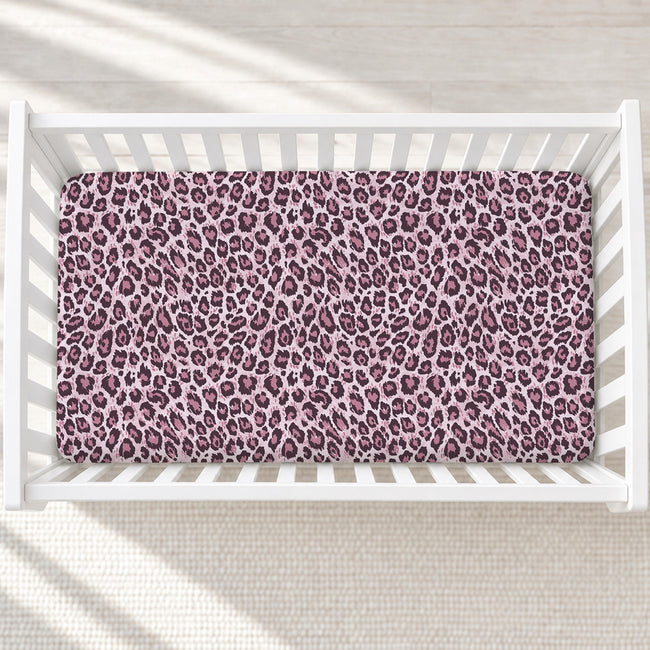 A rectangular baby crib mattress fitted with a pink and purple leopard print sheet, shown from a top-down view inside a white wooden crib.