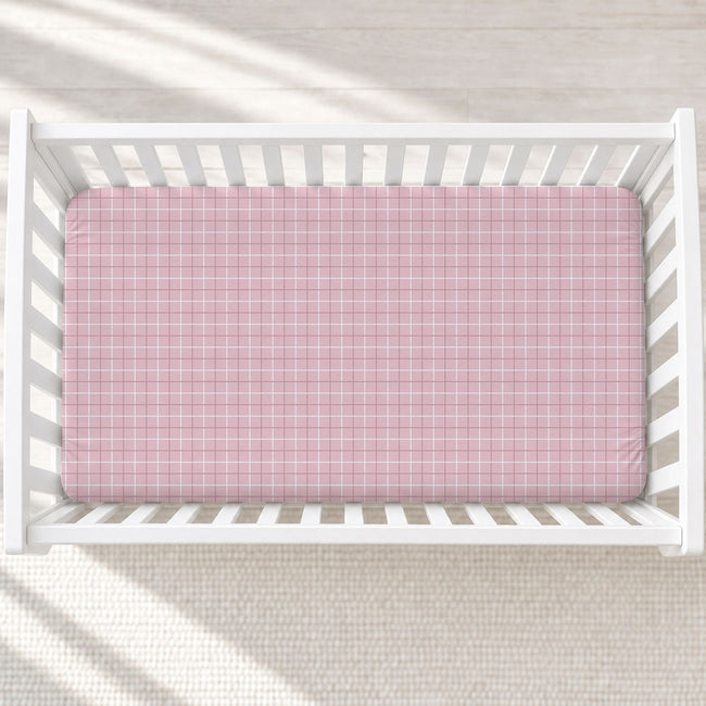 A pink and white plaid fitted sheet on a mattress inside a white wooden crib, shown from a top-down view on a light textured rug.