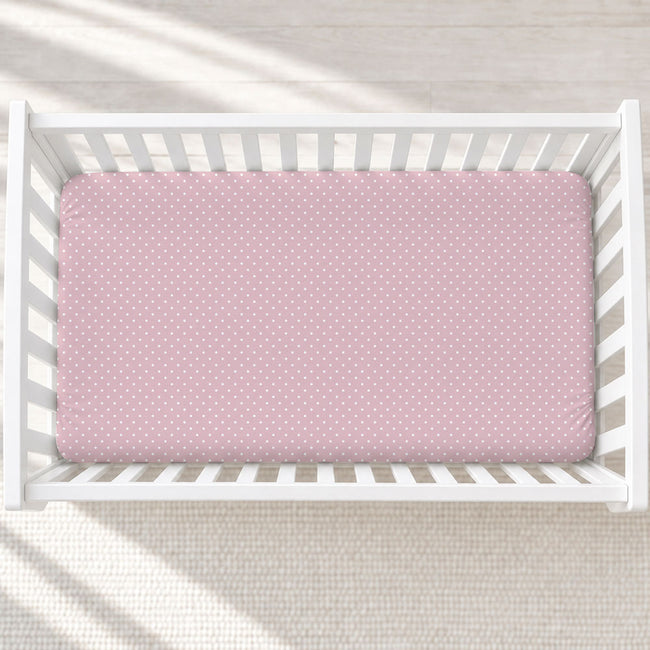 A pink fitted crib sheet with a white polka dot pattern, shown inside a white wooden baby crib on a light textured rug.
