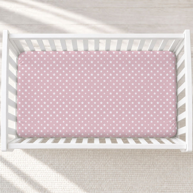 A pink fitted crib sheet with a white star pattern, shown inside a white wooden baby crib on a light textured rug.