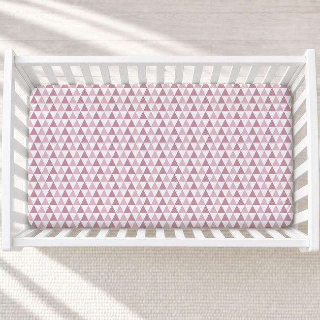 A top-down view of a white wooden crib with a pink and white geometric triangle patterned crib sheet, set against a light textured rug with soft window light shadows.