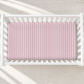 A pink mattress protector with thin white vertical stripes, shown inside a white wooden crib on a light textured rug.