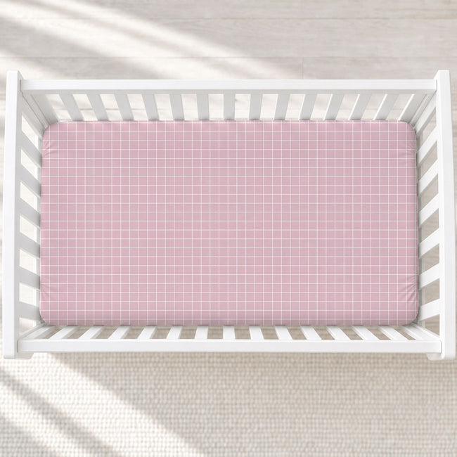 A pink fitted crib sheet with a white grid pattern, shown inside a white wooden baby crib from a top-down view.
