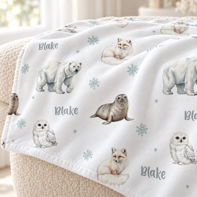 A white fleece throw blanket draped over a beige armchair, featuring watercolor illustrations of polar bears, seals, owls, foxes, and snowflakes, with the name 'Blake' printed in gray.