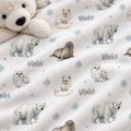 A close-up of a soft fleece blanket with a white polar bear and snowflake pattern, featuring watercolor illustrations of polar bears, seals, foxes, and owls, with the name 'Blake' printed in gray. A plush cream teddy bear is partially visible in the top left corner.