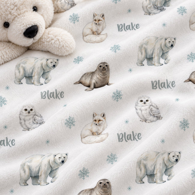 A close-up of a soft fleece blanket with a white polar bear and snowflake pattern, featuring watercolor illustrations of polar bears, seals, foxes, and owls, with the name 'Blake' printed in gray. A plush cream teddy bear is partially visible in the top left corner.