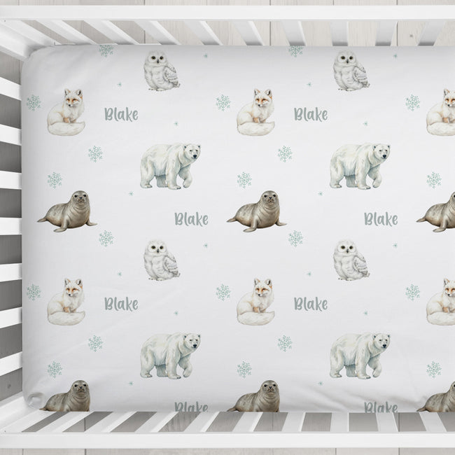 A personalized baby crib sheet with a repeating pattern of polar bears, seals, owls, and snowflakes, featuring the name 'Blake' in gray script. The sheet is displayed inside a white wooden crib.