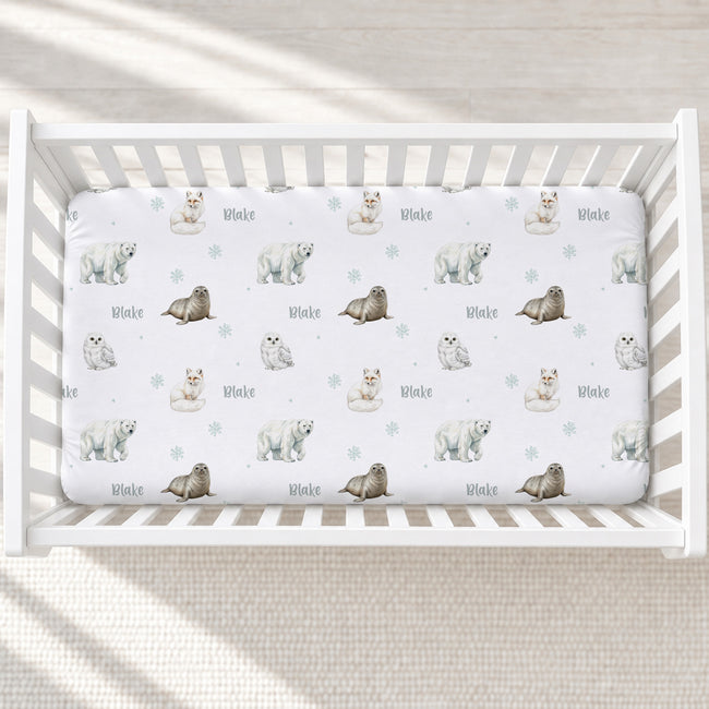 A white crib mattress fitted sheet with a repeating pattern of polar bears, seals, and the name 'Blake' in light blue-gray, shown inside a white wooden crib on a neutral textured floor.