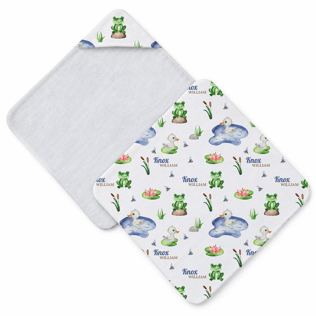 A white baby hooded towel with a whimsical pond and frog pattern, featuring green frogs, blue water, pink lotus flowers, and the personalized name 'Inox William' printed repeatedly. The reverse side is solid white.