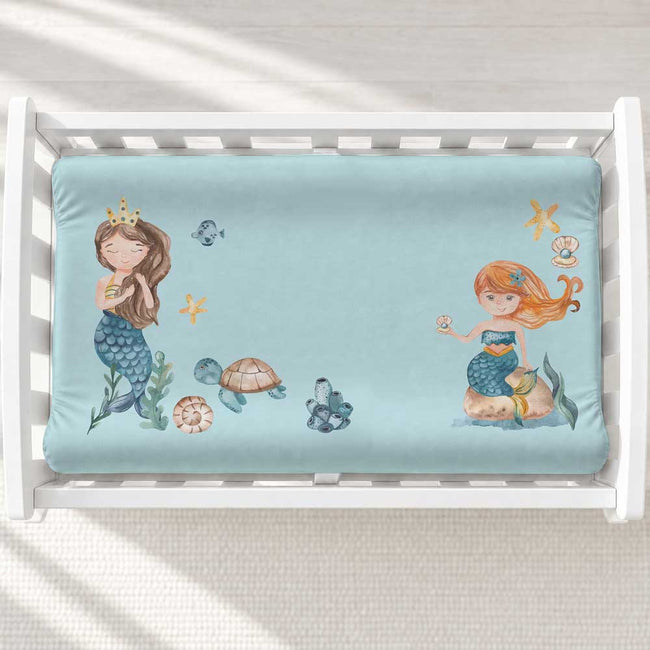 A light blue baby changing pad cover featuring a whimsical watercolor mermaid illustration, shown inside a white wooden crib on a textured cream rug.
