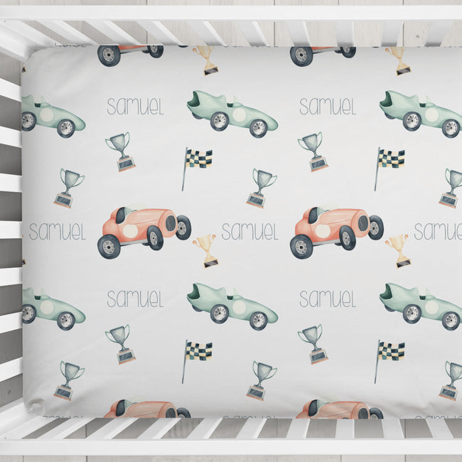 A baby crib bedding set featuring a white sheet with a repeating pattern of pastel race cars, trophies, checkered flags, and the name 'samuel' printed in gray.