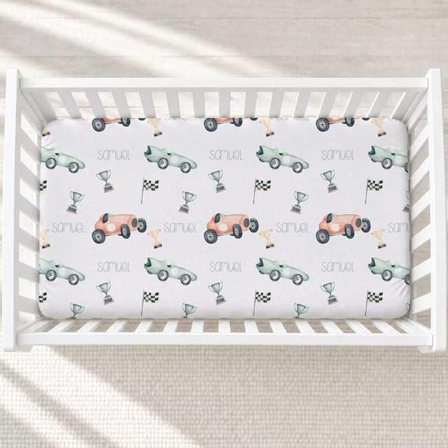 A white crib mattress fitted sheet with a race car pattern, personalized with the name 'Samuel' and decorated with retro cars, checkered flags, and trophies, shown in a baby crib.
