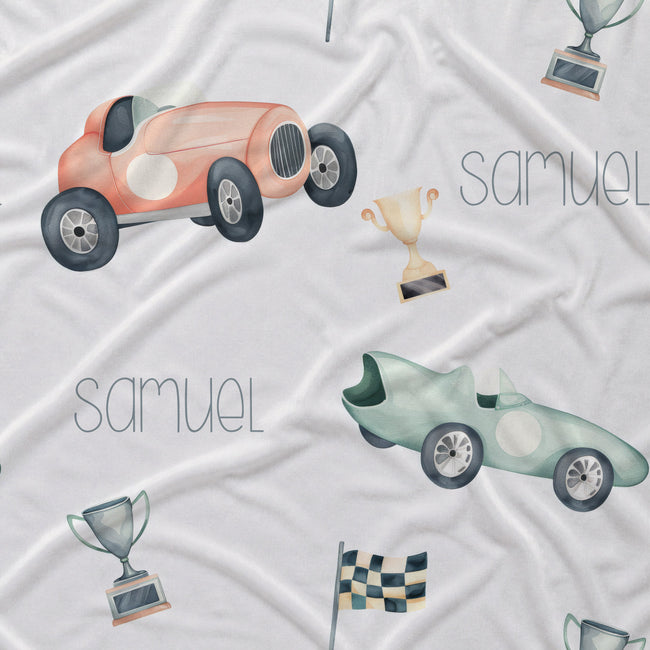 White fabric with a watercolor-style print of vintage racing cars, trophies, checkered flags, and the name 'samuel' in a thin gray font.