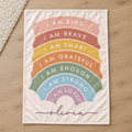 Personalized Olivia blanket lying on a wood floor, featuring a pastel rainbow with seven positive affirmations and a mauve script name on cream.