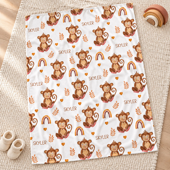 A white blanket with a repeating cartoon monkey print, rainbow motifs, and the name 'SKYLER' scattered across it, styled on a woven rug with small slippers and a wooden toy nearby.