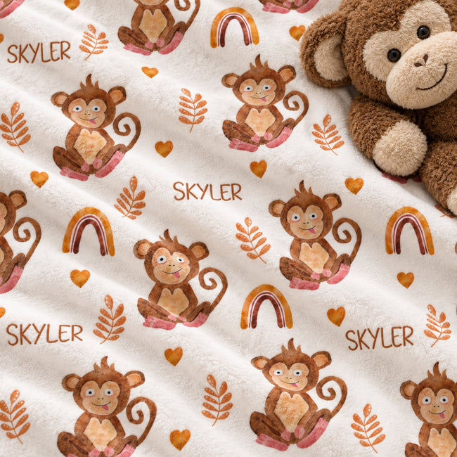 A close-up of a plush blanket with a repeating pattern of brown cartoon monkeys, orange leaves, small rainbows, and the name 'SKYLER' printed in brown. A brown plush monkey toy is partially visible in the top-right corner on a white background.