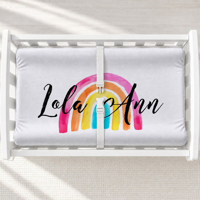 A white changing table with a personalized changing pad cover featuring a colorful watercolor rainbow and the personalized name 'Lola Ann' in black script, shown in a white crib with a safety strap.