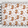 A crib fitted sheet featuring a repeating pattern of cartoon brown monkeys, rainbows, leaves, and the name 'SKYLER' printed in brown on a white background, shown inside a white crib.