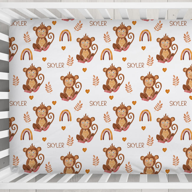 A crib fitted sheet featuring a repeating pattern of cartoon brown monkeys, rainbows, leaves, and the name 'SKYLER' printed in brown on a white background, shown inside a white crib.