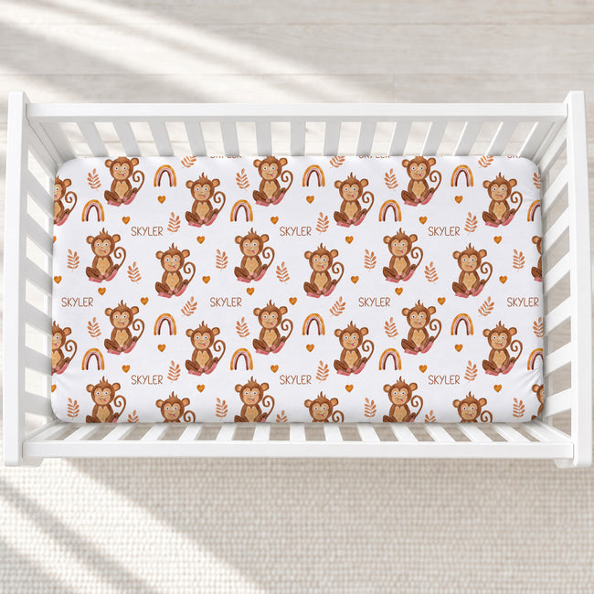 A white crib mattress fitted sheet with a repeating pattern of brown cartoon monkeys, rainbows, leaves, and the word 'SKYLER', shown inside a white crib on a light textured floor.
