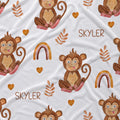 A white fabric blanket or towel featuring a playful pattern of cartoon baby monkeys, brown hearts, leaf motifs, and rainbows, with the name 'SKYLER' printed in brown text.