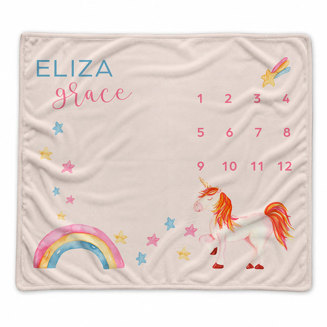 A personalized baby blanket featuring a white unicorn with orange and pink details, a pastel rainbow, scattered stars, and the name 'ELIZA grace' alongside a numbered calendar grid, all on a soft beige background.