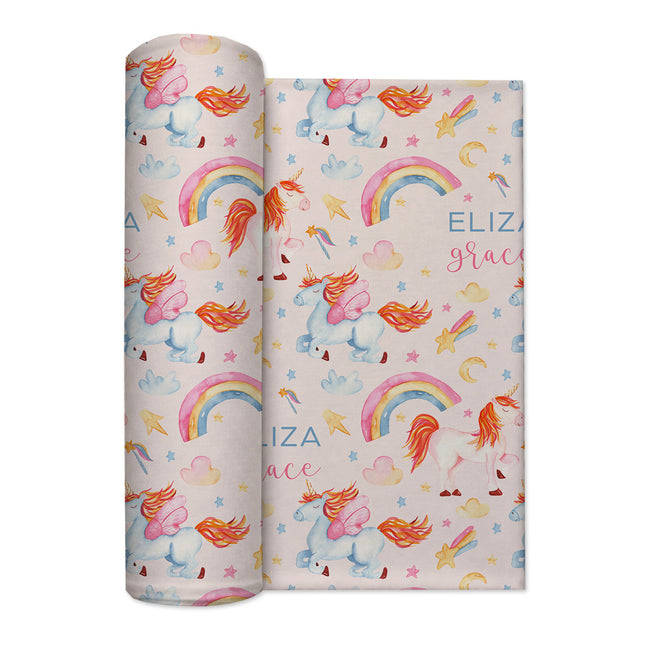A rolled personalized swaddle blanket and a flat sheet displaying a whimsical unicorn pattern. The design features pastel blue unicorns with orange manesues, pink bows, colorful rainbows, stars, and hearts on a light cream background.