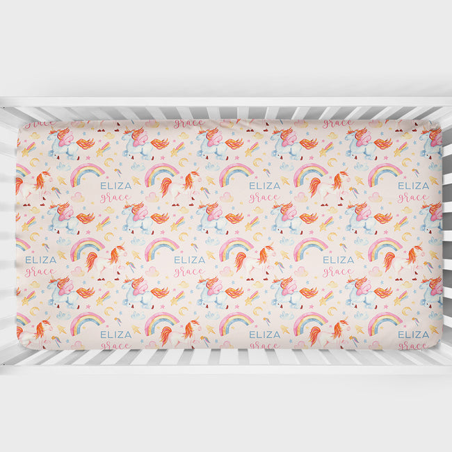A crib mattress fitted sheet with a whimsical unicorn and rainbow pattern, shown inside a white wooden crib. The sheet features a light pink background with orange, pink, and blue unicorns, pastel rainbow arches, and the text 'ELIZA grace' repeated throughout.