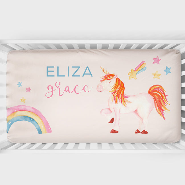 Personalized baby crib sheet with a pastel unicorn, rainbow, and star illustrations, featuring the names 'ELIZA' and 'grace' in blue and pink lettering on a cream background.