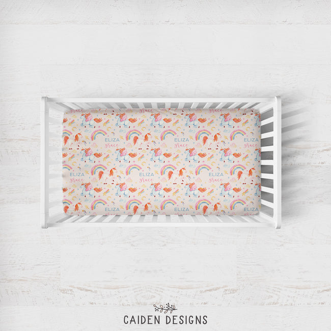 A white wooden baby crib with a colorful printed mattress protector featuring unicorns, rainbows, and the name 'ELIZA' repeated, shown on a light whitewashed wood background. The brand name 'CAIDEN DESIGNS' is printed at the bottom.