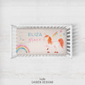 A personalized baby bedding set displayed inside a white wooden crib, featuring a cream-colored pillowcase with a whimsical unicorn, rainbow, and star design, along with the custom names 'ELIZA Grace'.