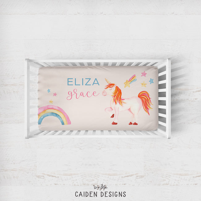 A personalized baby bedding set displayed inside a white wooden crib, featuring a cream-colored pillowcase with a whimsical unicorn, rainbow, and star design, along with the custom names 'ELIZA Grace'.