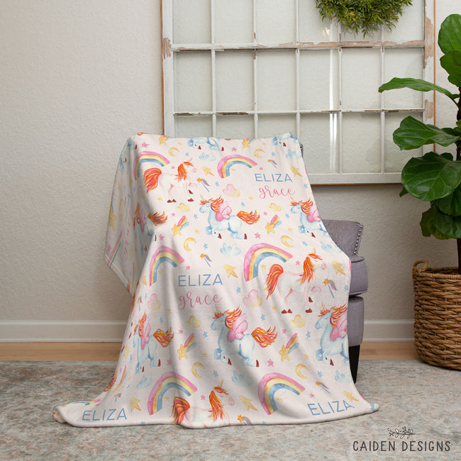 A pastel fleece throw blanket with a whimsical unicorn and rainbow print draped over a gray armchair in a styled room setting. The blanket features the names 'ELIZA' and 'GRACE' printed in blue and pink script, along with colorful unicorns, rainbows, and stars.