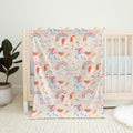 A personalized baby blanket with a colorful unicorn and rainbow pattern, draped over a light wood crib in a nursery setting.