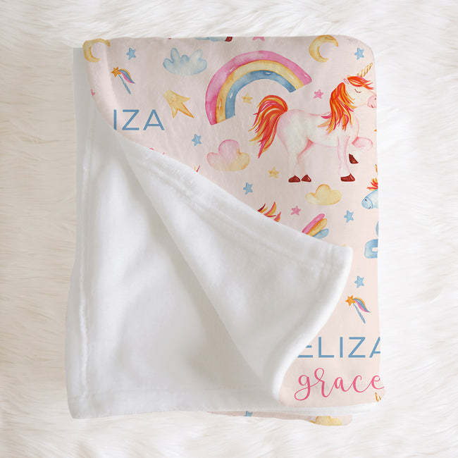 A personalized baby blanket with a white sherpa backing and a printed front featuring a unicorn, rainbow, stars, and clouds on a light pink background. The name 'ELIZABETH' is printed in blue and pink lettering.
