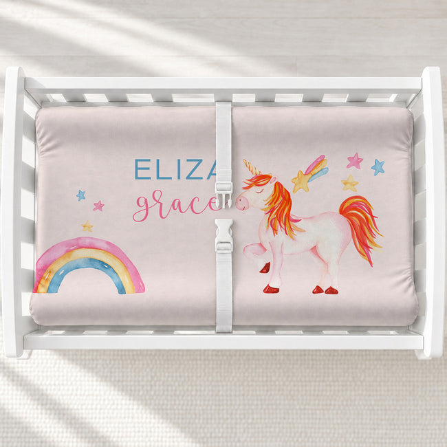 A white changing table with a personalized changing pad cover featuring a white unicorn, rainbow, and stars, featuring the name 'ELIZABETH GRACIE' in blue and pink lettering. 