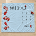 A personalized baby blanket laid flat on a light wood floor. The blanket is light blue with black text reading 'NOAH SPENCER' and a 12-month countdown grid, decorated with red fire trucks, hydrants, and a firefighter helmet.