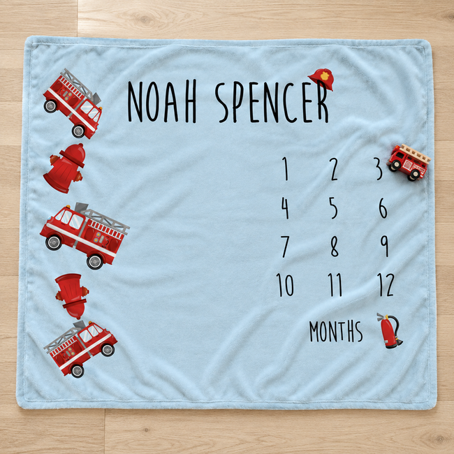 A personalized baby blanket laid flat on a light wood floor. The blanket is light blue with black text reading 'NOAH SPENCER' and a 12-month countdown grid, decorated with red fire trucks, hydrants, and a firefighter helmet.
