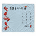 A personalized baby blanket in light blue with a fire department theme. The blanket features the name 'NOAH SPENCER' in black text, a 12-month countdown grid, and illustrations of red fire trucks, hydrants, and a firefighter helmet.