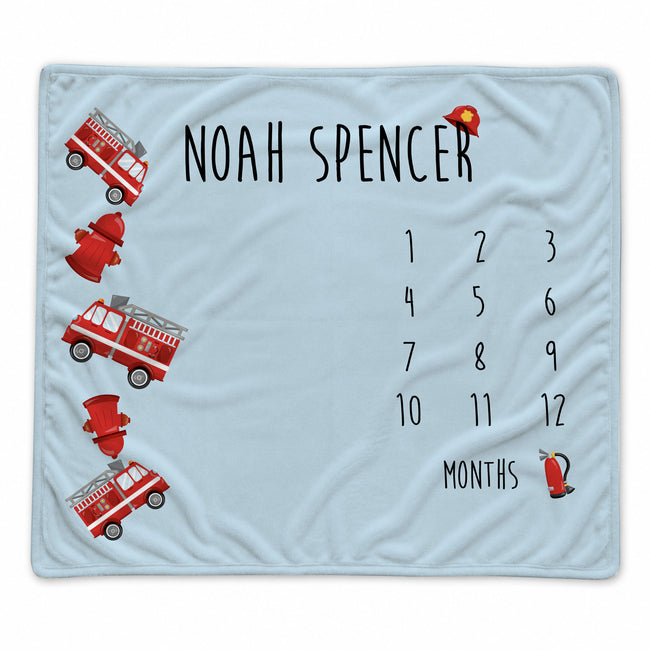 A personalized baby blanket in light blue with a fire department theme. The blanket features the name 'NOAH SPENCER' in black text, a 12-month countdown grid, and illustrations of red fire trucks, hydrants, and a firefighter helmet.