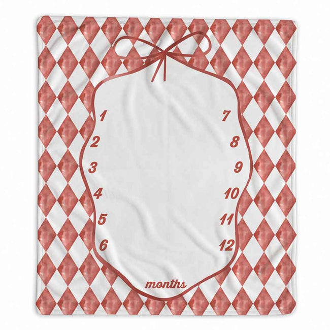 A square baby milestone blanket featuring a red and white harlequin diamond pattern border, a central white oval space outlined in red with the words 'months' and numbers 1 through 12, and a red bow illustration at the top.