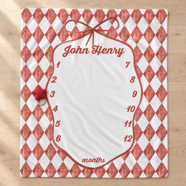 A personalized monthly milestone blanket with a red and white harlequin diamond pattern border, a large white writing area, and a small red star ornament hanging from a metal ring on the left side, shown on a light wood floor.