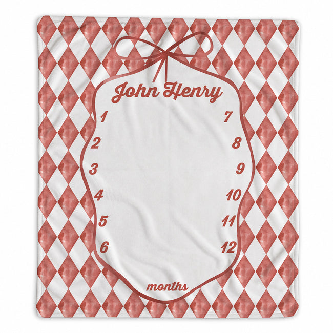 A personalized baby milestone chart print featuring a red and white harlequin diamond pattern border, a central white oval space with a red bow illustration, and the name 'John Henry' at the top. The chart includes numbered slots from 1 to 12 for months and is shown flat against a white background.