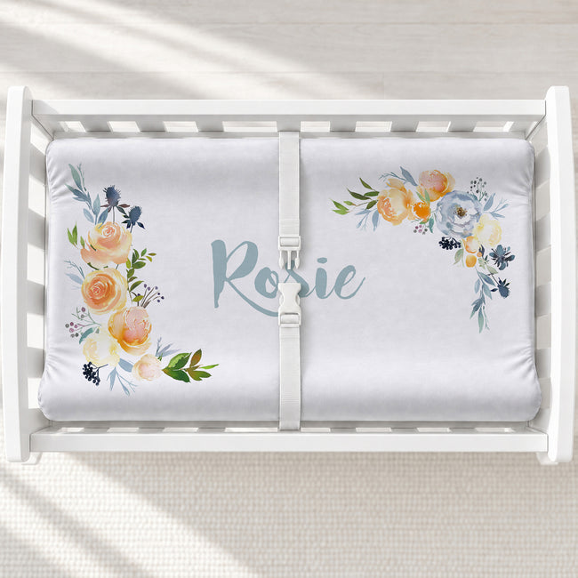 A white changing table with a personalized changing pad cover featuring a floral bumper sticker personalized with the name 'Rosie' in light blue script. The floral design features peach, yellow, and blue watercolor flowers with green leaves.