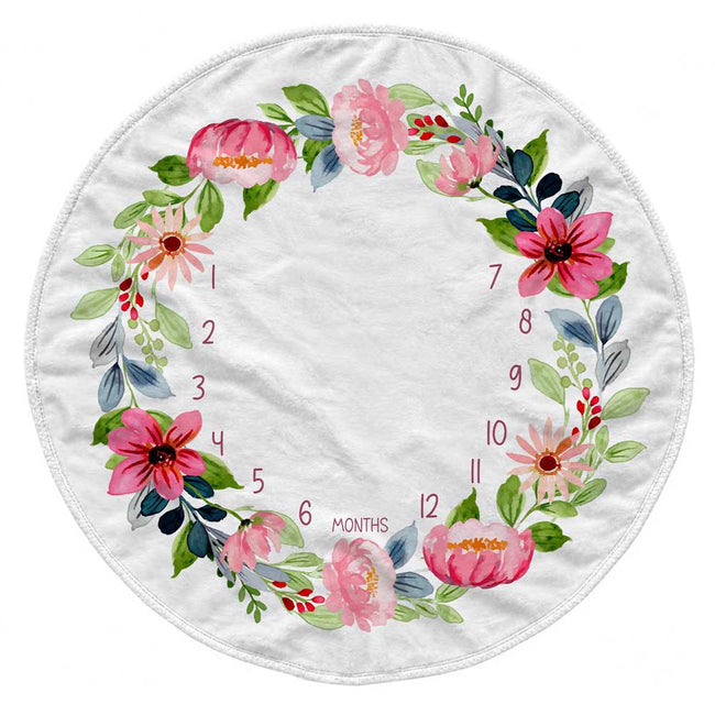 A round white baby blanket featuring a watercolor floral wreath design in pink, green, and blue, with printed numbers 1 through 12 and the word 'MONTHS' arranged in the center.