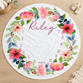 A round white baby milestone blanket with a colorful floral wreath and the name 'Riley' in script, shown on a light wood floor with small props around it.