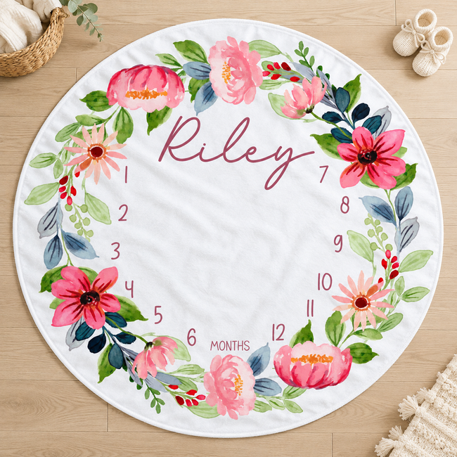 A round white baby milestone blanket with a colorful floral wreath and the name 'Riley' in script, shown on a light wood floor with small props around it.