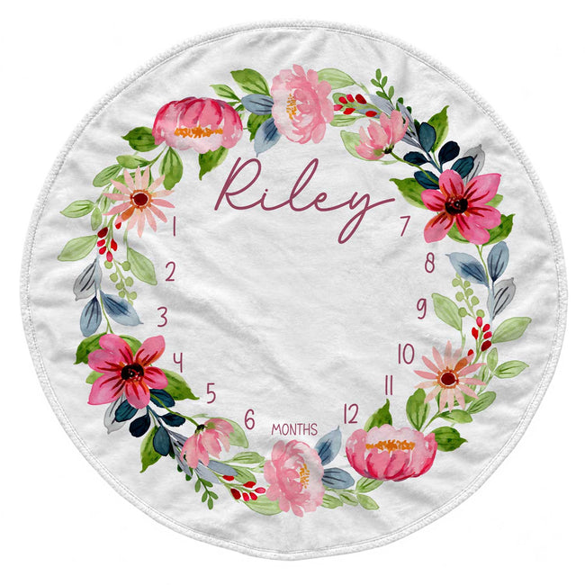 A round white baby blanket featuring a colorful watercolor floral wreath with the name 'Riley' in script and numbers 1 through 12 arranged around the border for monthly milestone tracking.