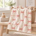 An off-white baby blanket with a red bow and chain pattern and the word 'lively' embroidered in red, draped over a woven basket next to a wooden bench in a bright nursery setting.