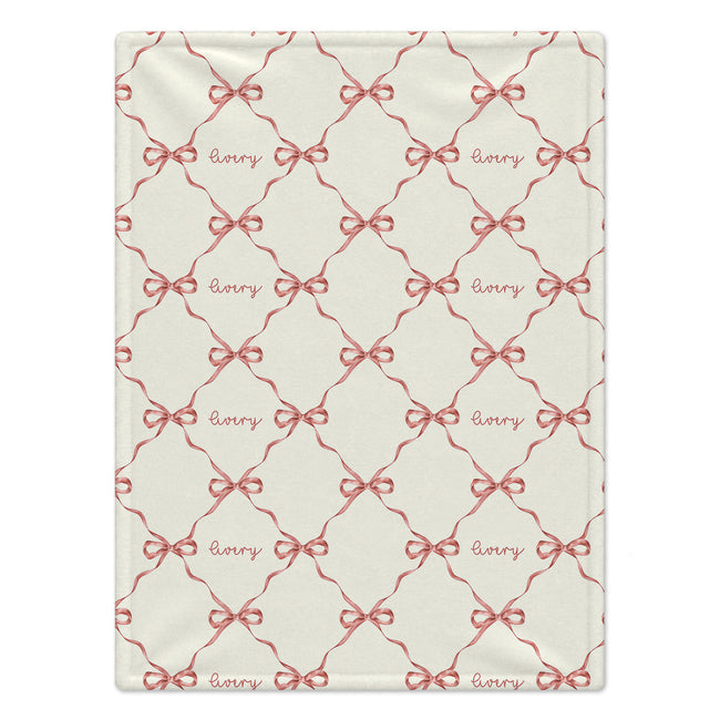 A rectangular cream-colored blanket featuring a repeating pattern of red ribbon bows and the word 'Avery' in cursive script.